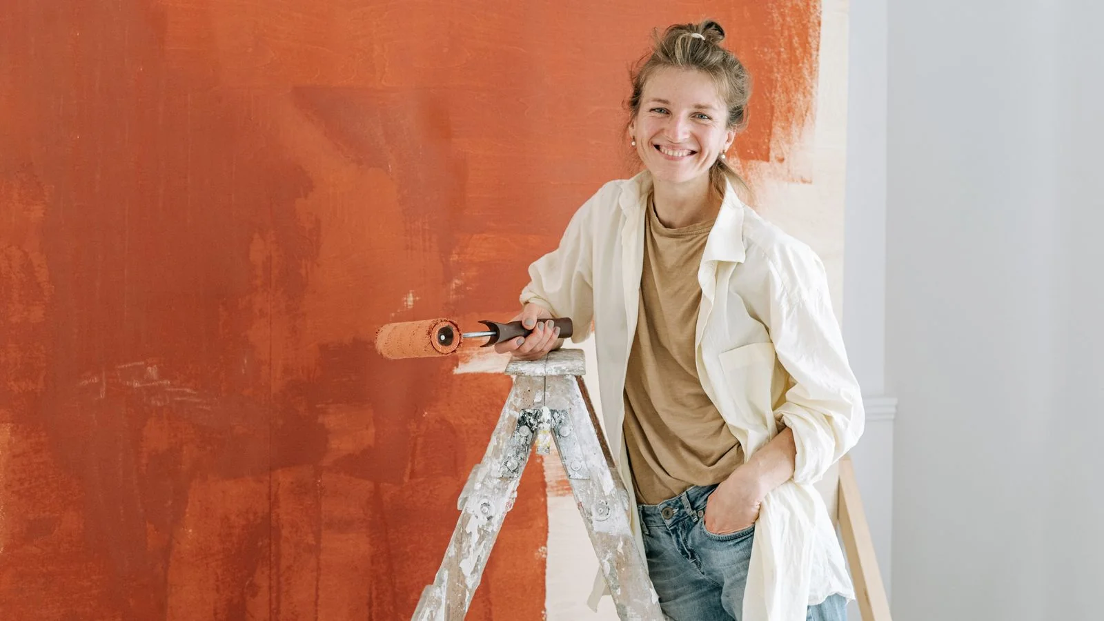 A painter on a ladder rolling orange paint across an interior wall.