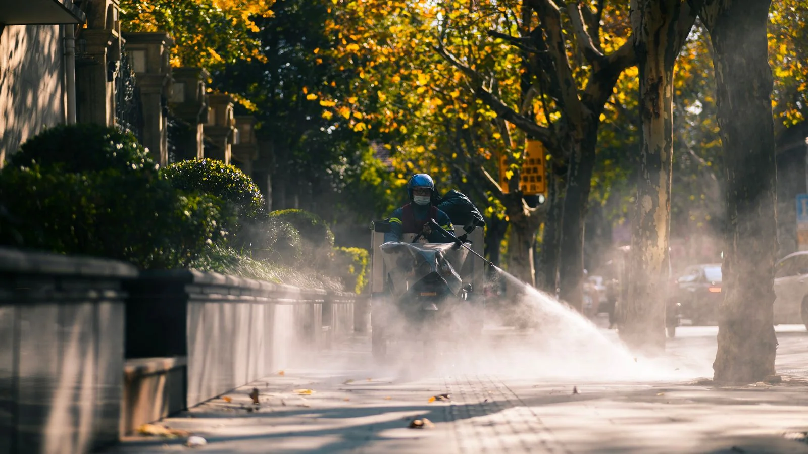 A city worker spraying down a public sidewalk on a sunny day, the community-facing side of the work.
