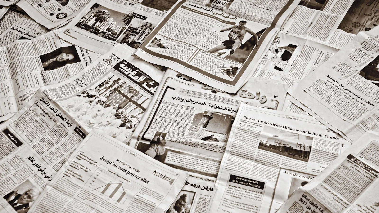 Stack of newspapers in multiple languages including Arabic and French, spread across a table