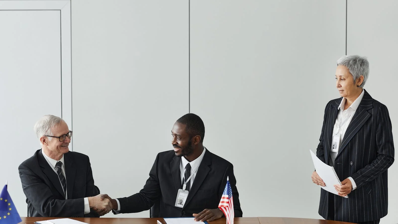Two businessmen shaking hands across a conference table, with flags from different countries between them