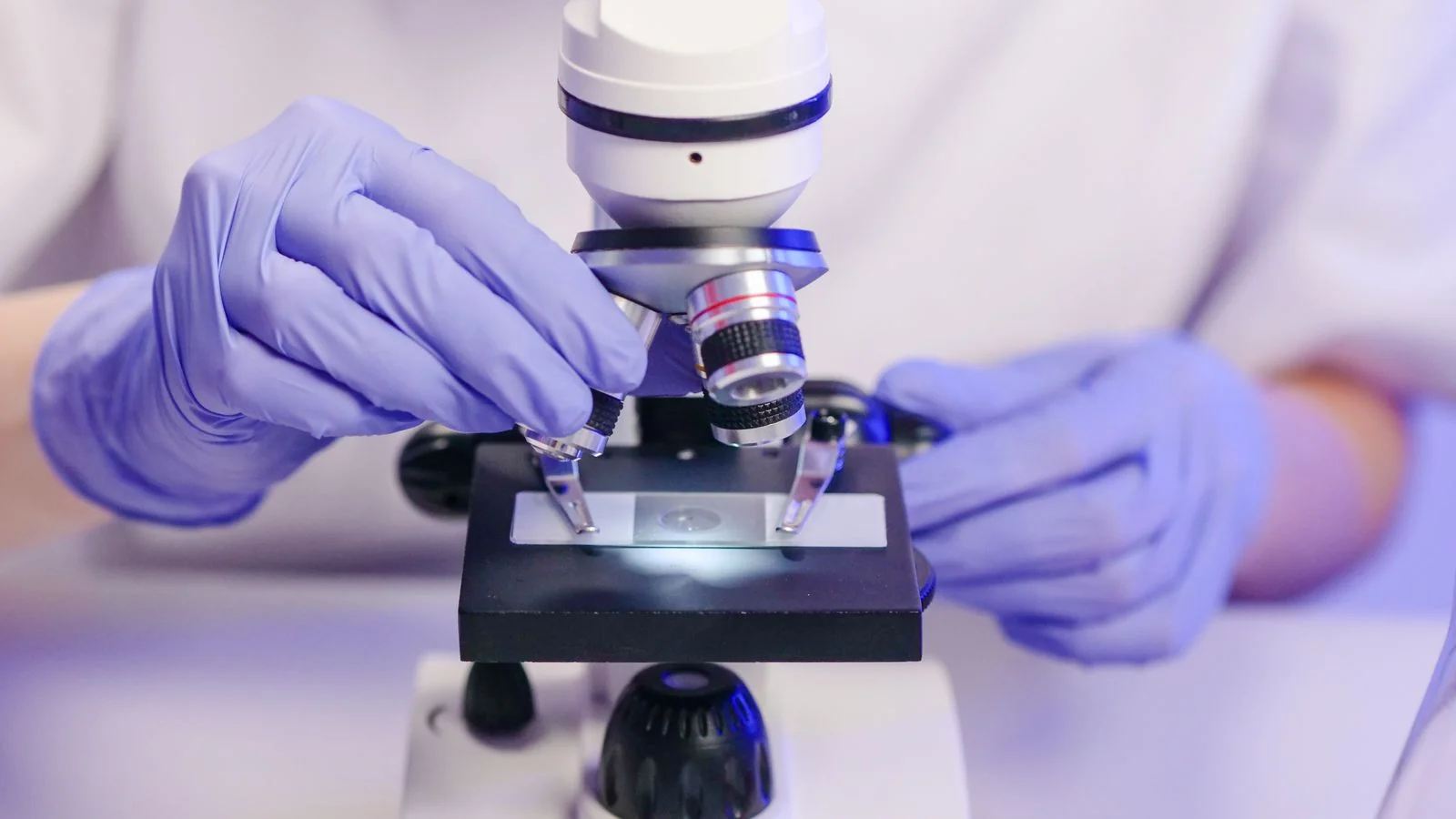 A gloved scientist adjusts a microscope while analyzing a sample
