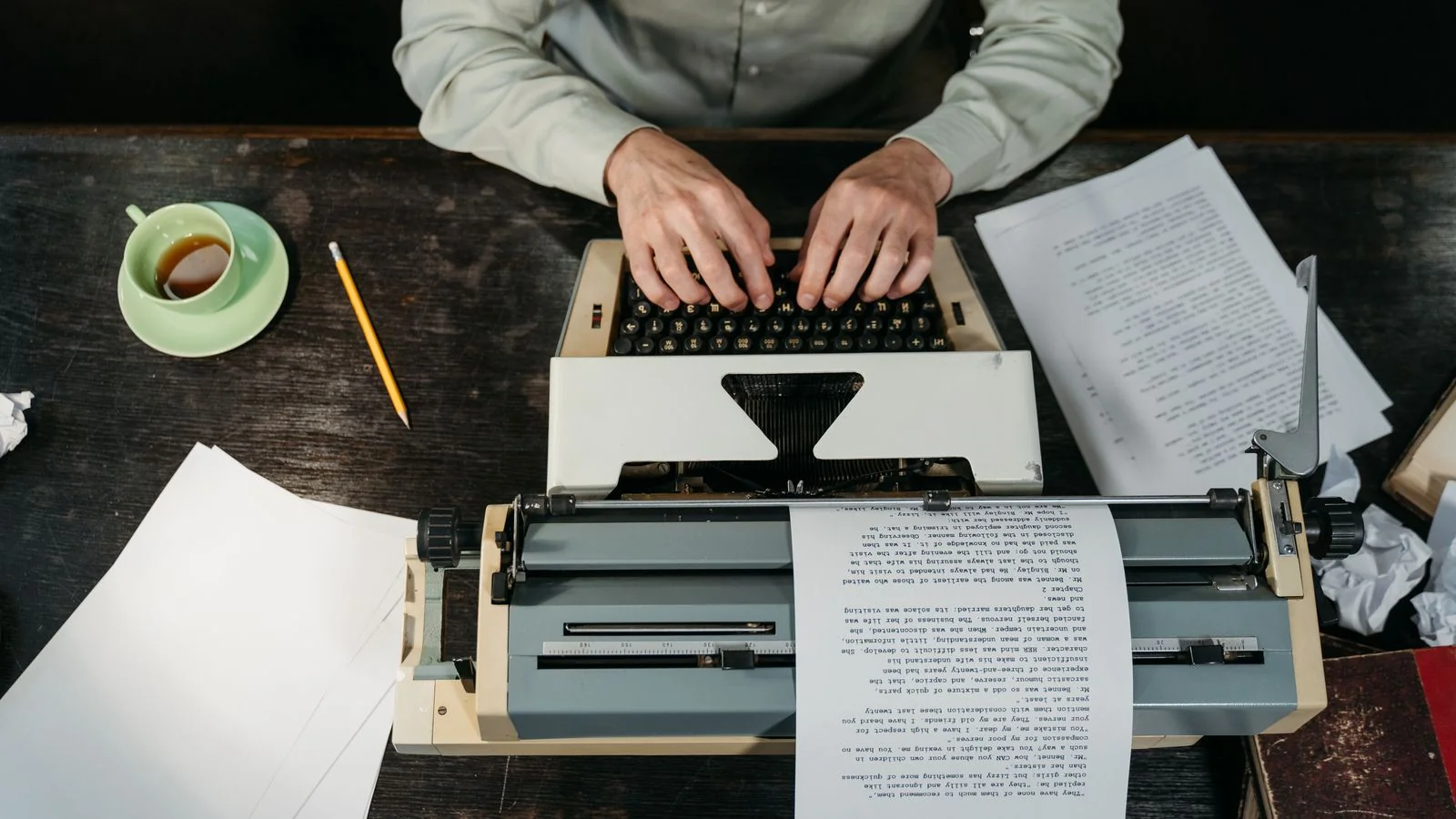 Hands typing a draft on a typewriter beside loose papers and a coffee cup on a desk
