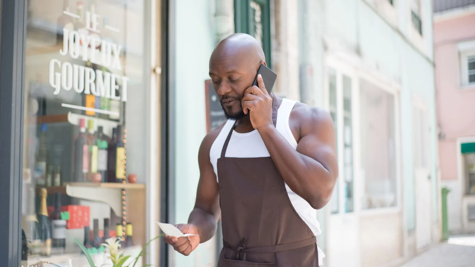 Man in an apron taking a phone call and reading a note outside his small shop