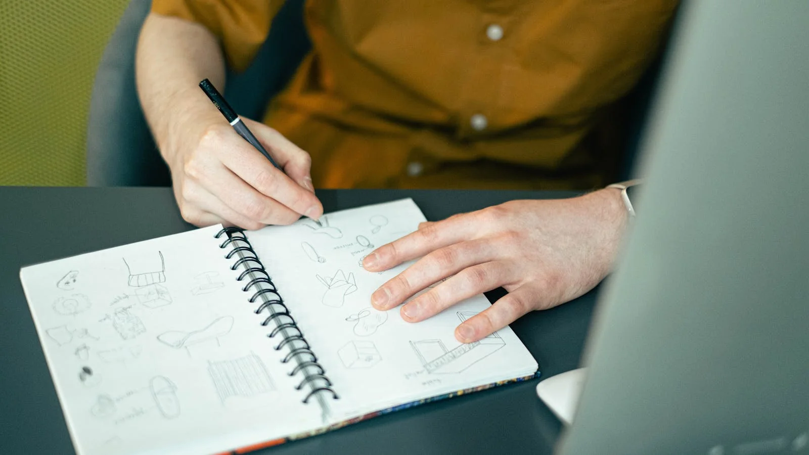 A designer's hands sketching ideas in a spiral notebook on a desk, pencils and reference notes arranged nearby during a naming workshop.