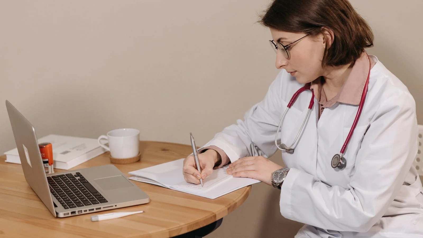 Doctor with a stethoscope writing notes at a desk beside a laptop