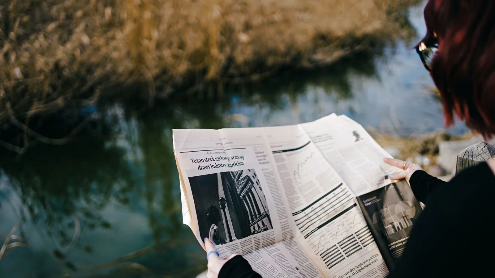 Person reading a newspaper outdoors beside a calm pond on a quiet morning