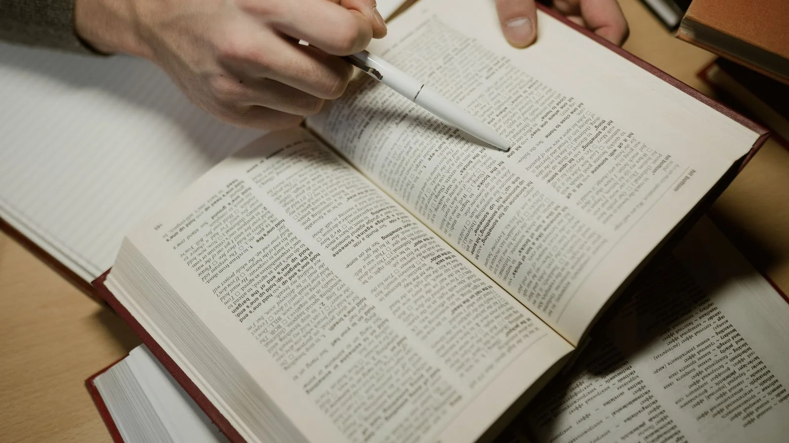 A hand holding a pen while studying an open book, the close reading Perplexity does to find sources worth citing