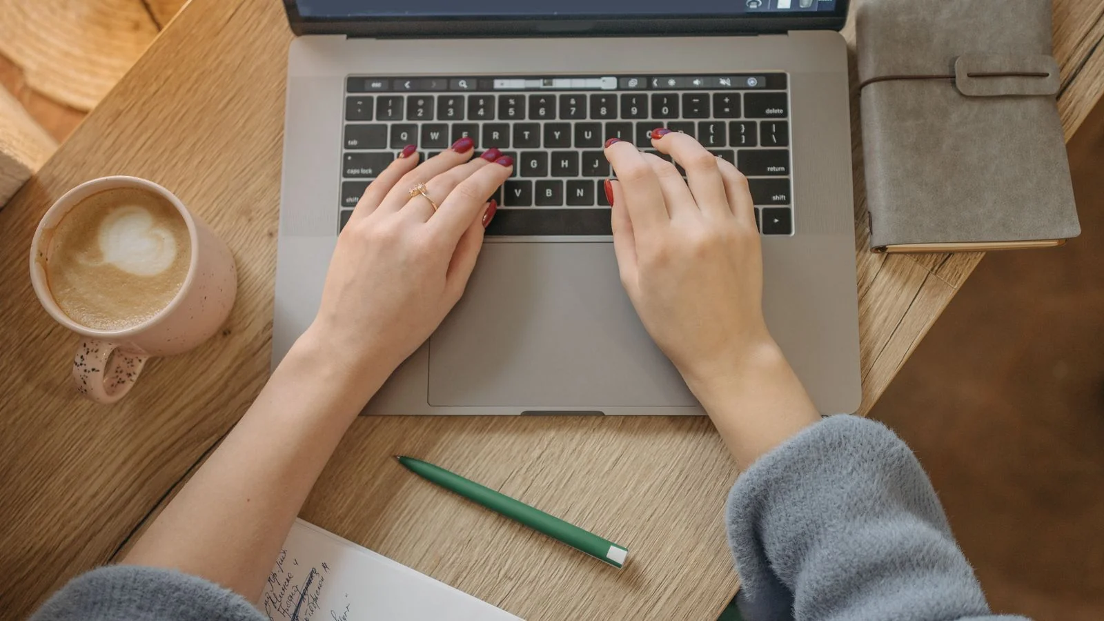 Top-down shot of a person typing on a laptop beside a notebook and pen on a wooden table