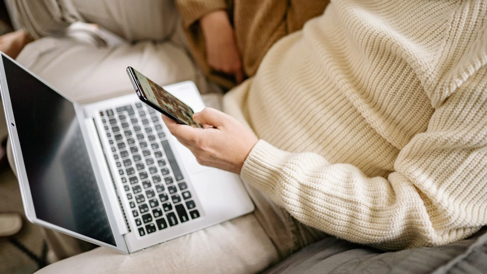 A person in a sweater multitasking with a smartphone and laptop indoors