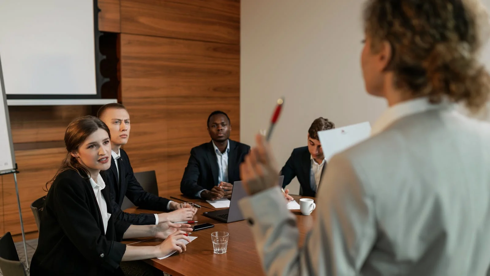 A businesswoman in a beige suit standing in a meeting room holding a pen, mid-conversation during a quarterly client review.