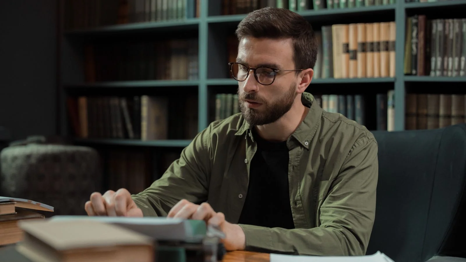 A young man in glasses typing at a vintage typewriter in a library, working through a piece one deliberate line at a time