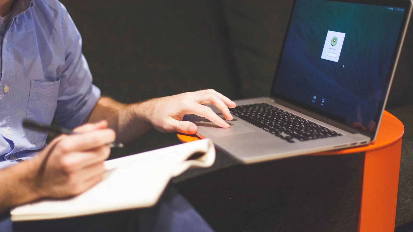 Man writing notes beside an open laptop, drafting the single sentence a company bio is built on