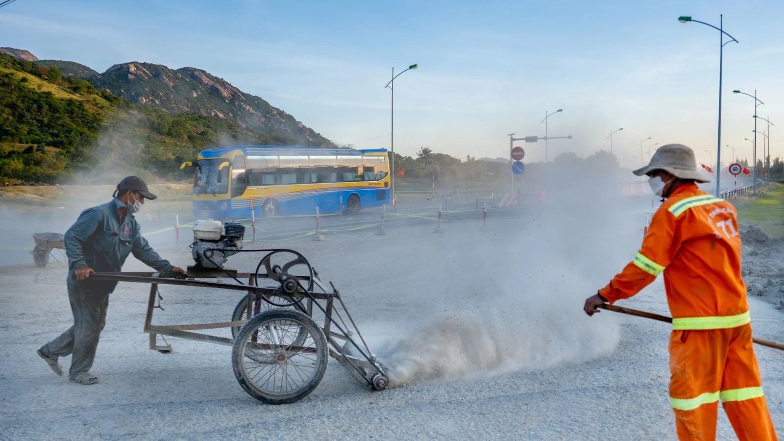 Two workers in protective gear running machinery on a dusty road as a bus passes