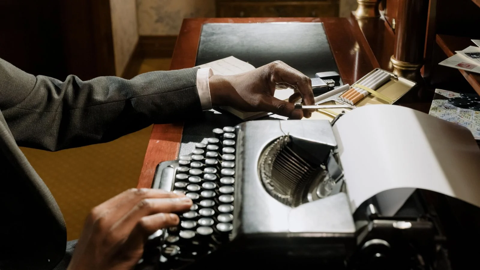 A journalist typing at a newsroom desk with notes and a monitor.