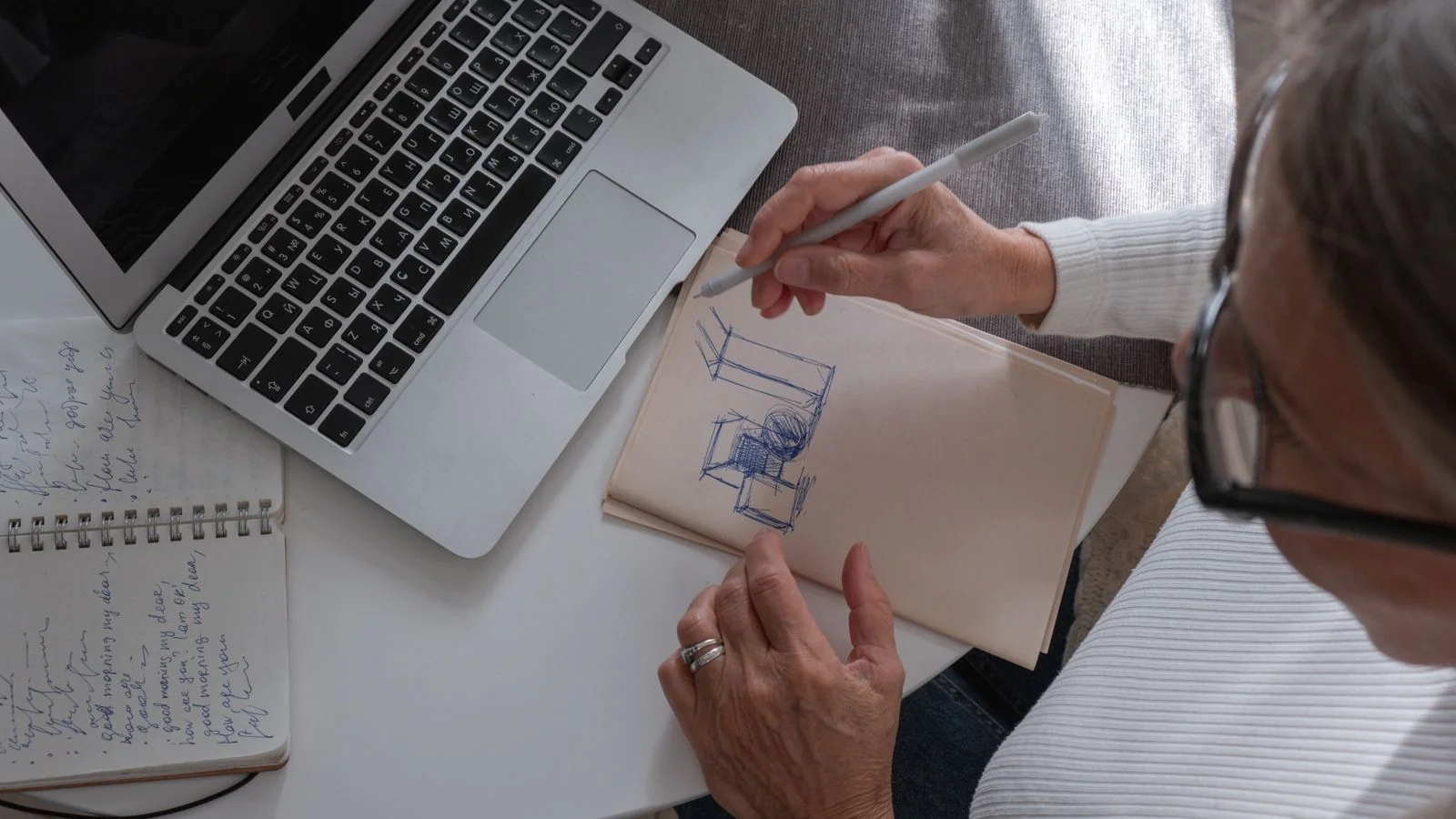 A person sketching notes at a home desk beside a laptop, drafting a release.