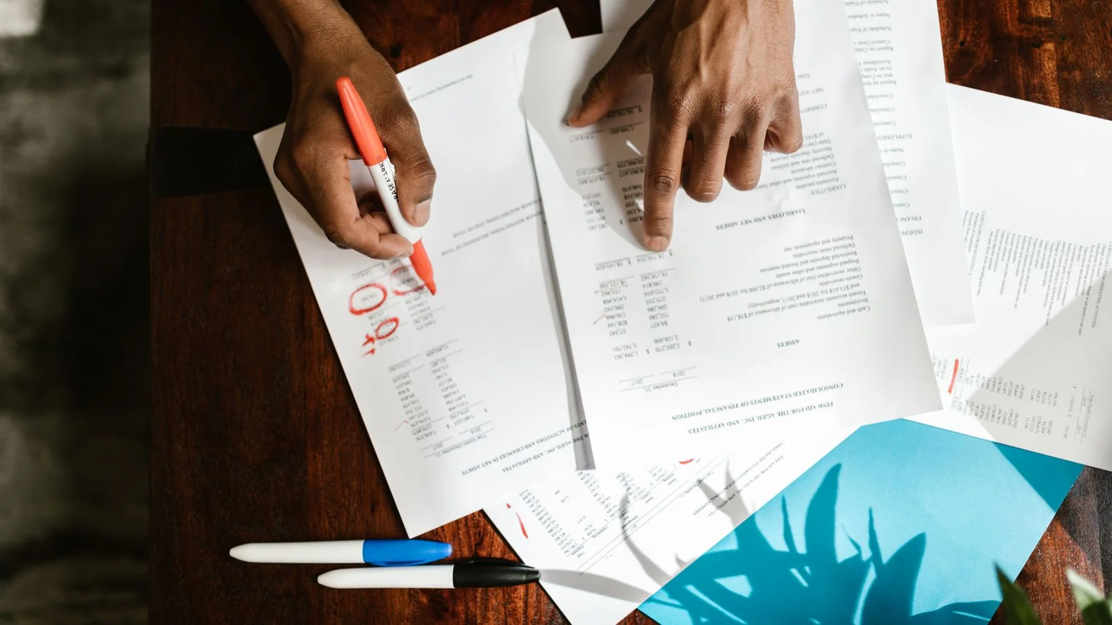 Hands mark up a printed report with a red marker, circling figures at a wooden desk.
