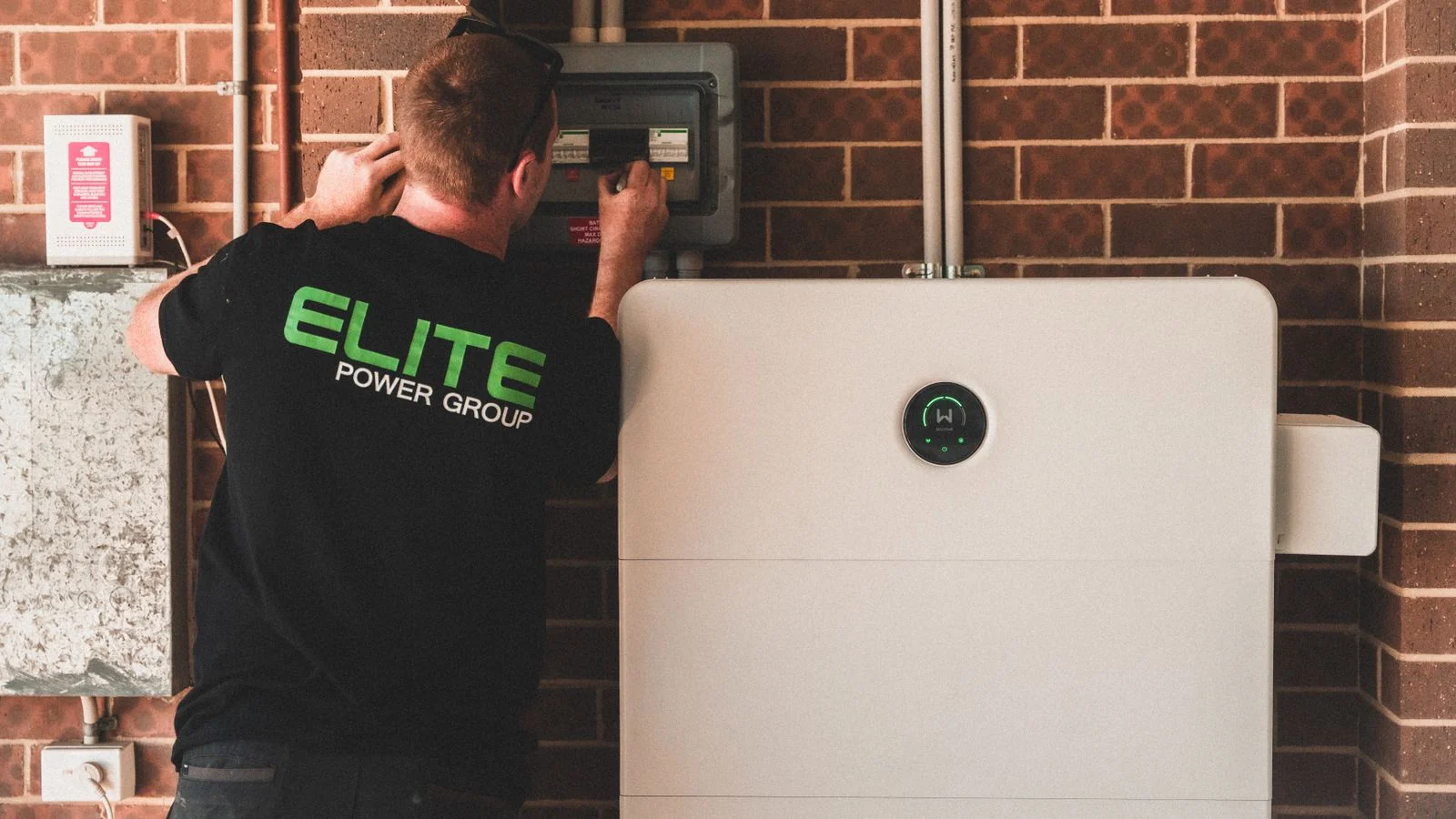 An inspector examines an electrical panel on a brick wall beside a home battery unit.