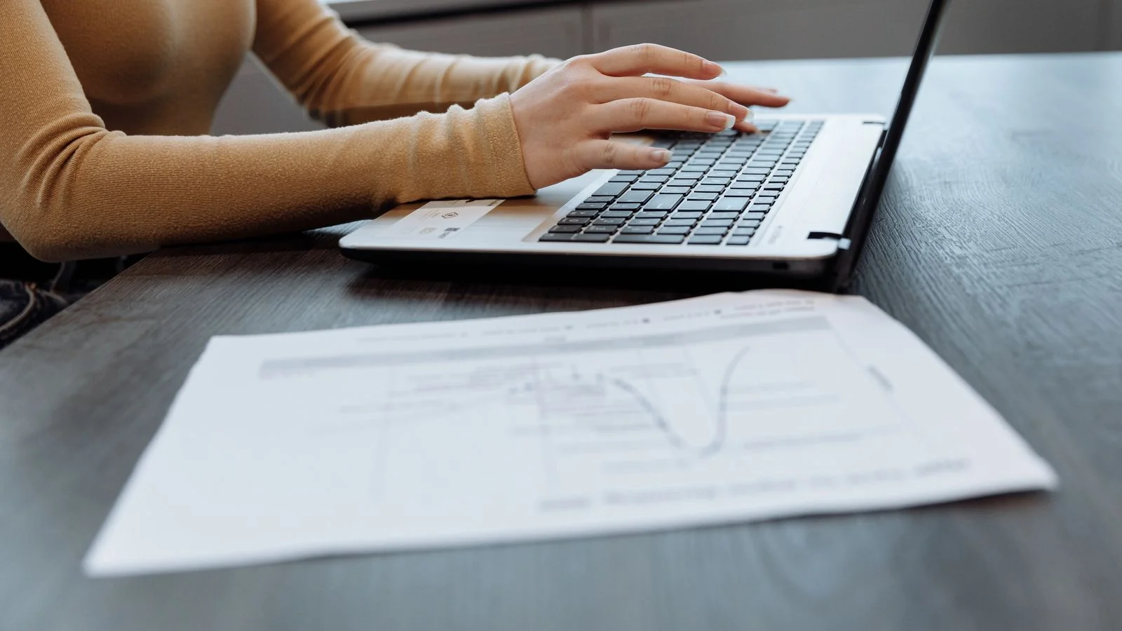 A woman in a tan sweater types a document on a laptop beside a printed chart at a desk.