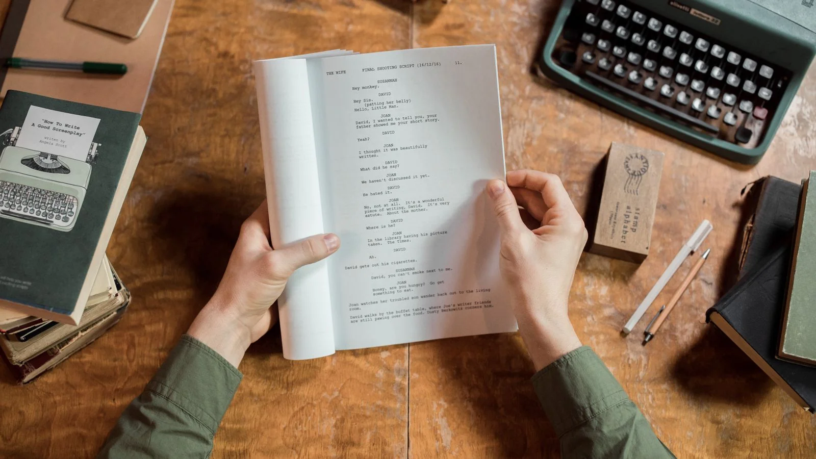 Hands holding a typed script page at a desk with a typewriter and books