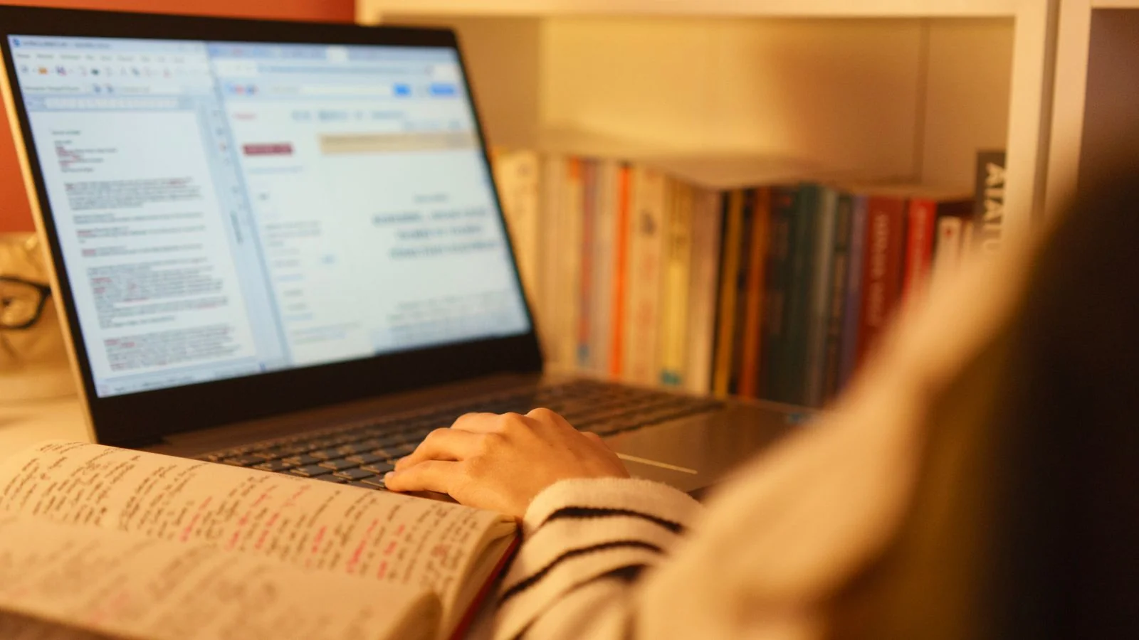 Person studying with a laptop and an open book in a home office lined with shelves, drafting an essay