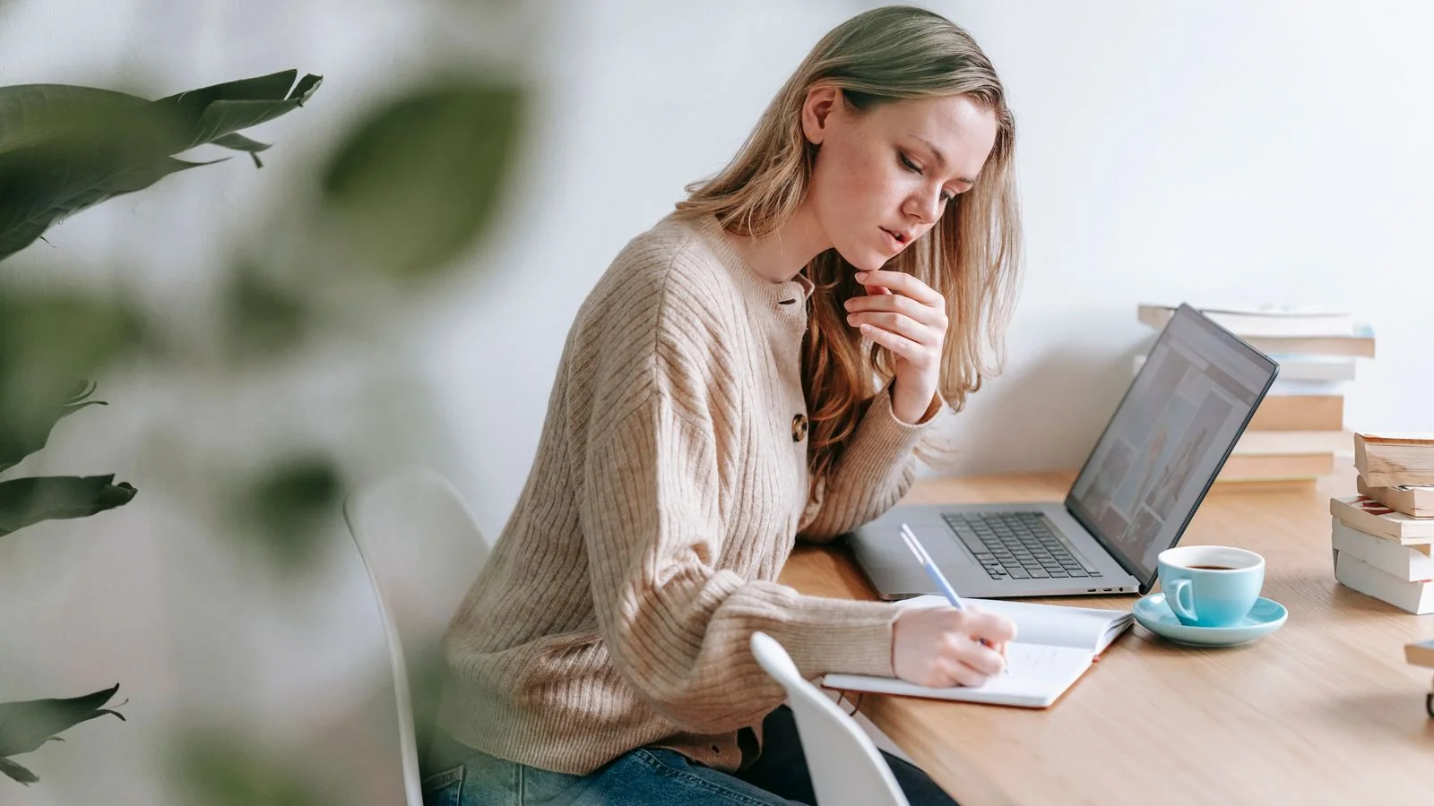 Woman writing notes in a notebook beside her laptop at home, building a public body of work