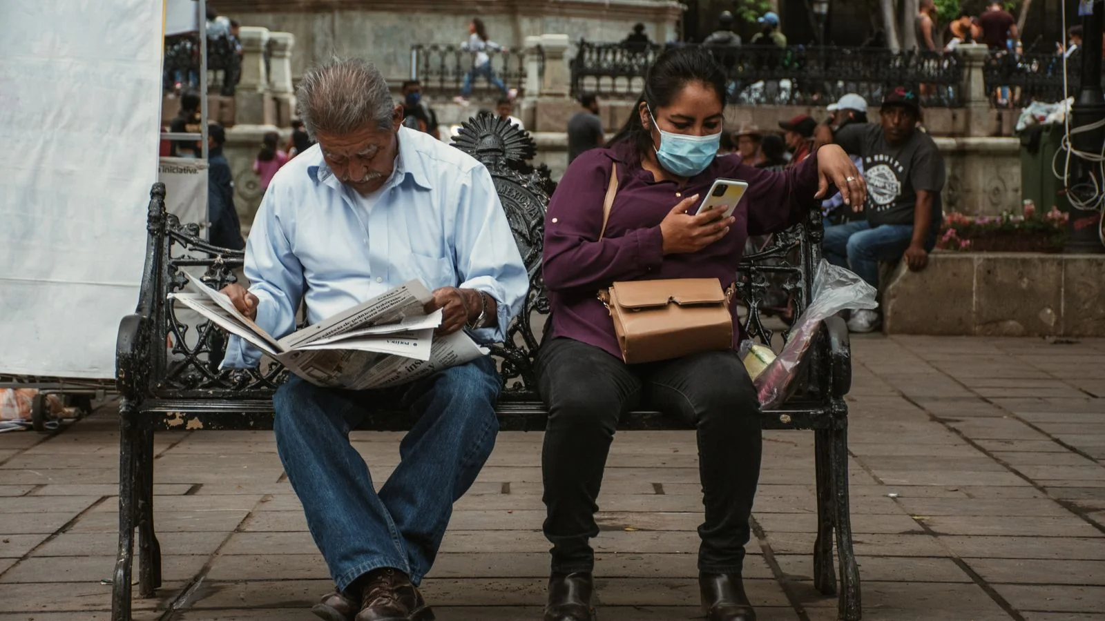 Man reading a newspaper beside a woman on her phone on a park bench
