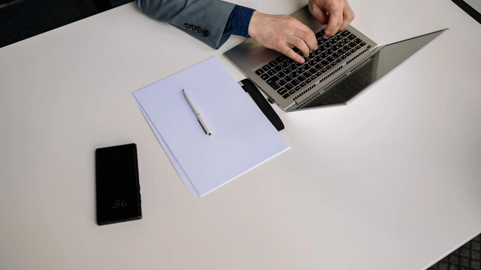 Businessperson typing on a laptop at a tidy white desk with phone and notes, the daily work behind a LinkedIn presence