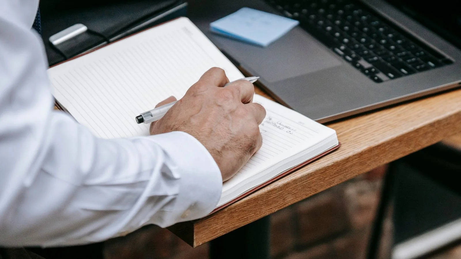 A person taking handwritten notes beside a laptop while preparing for an interview
