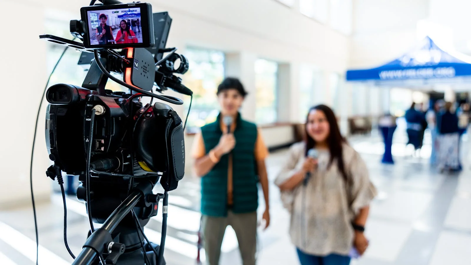 A professional video camera captures two interviewees during an indoor interview.