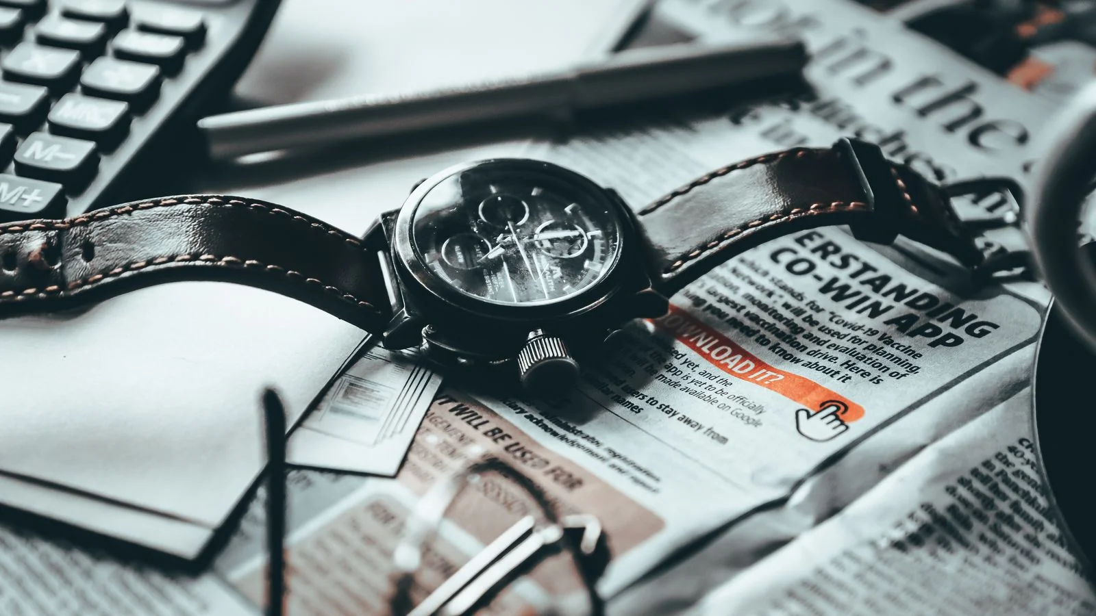 Wristwatch resting on a newspaper beside papers, a calculator, pen and eyeglasses on a table