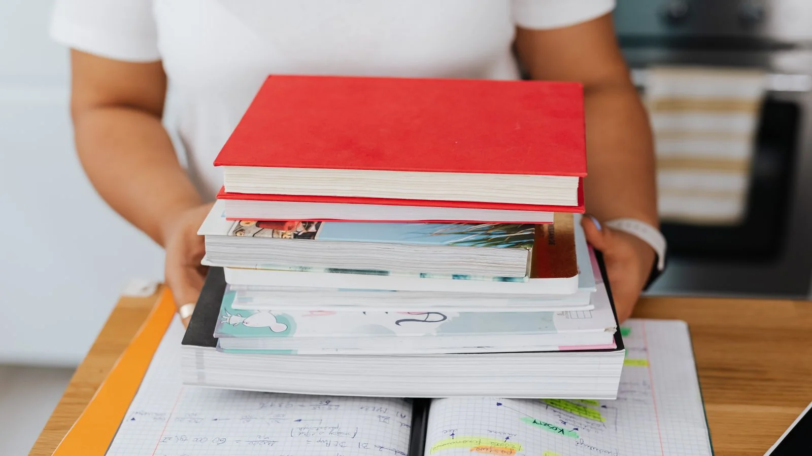 A person holding a stack of colorful books over an open notebook on a desk