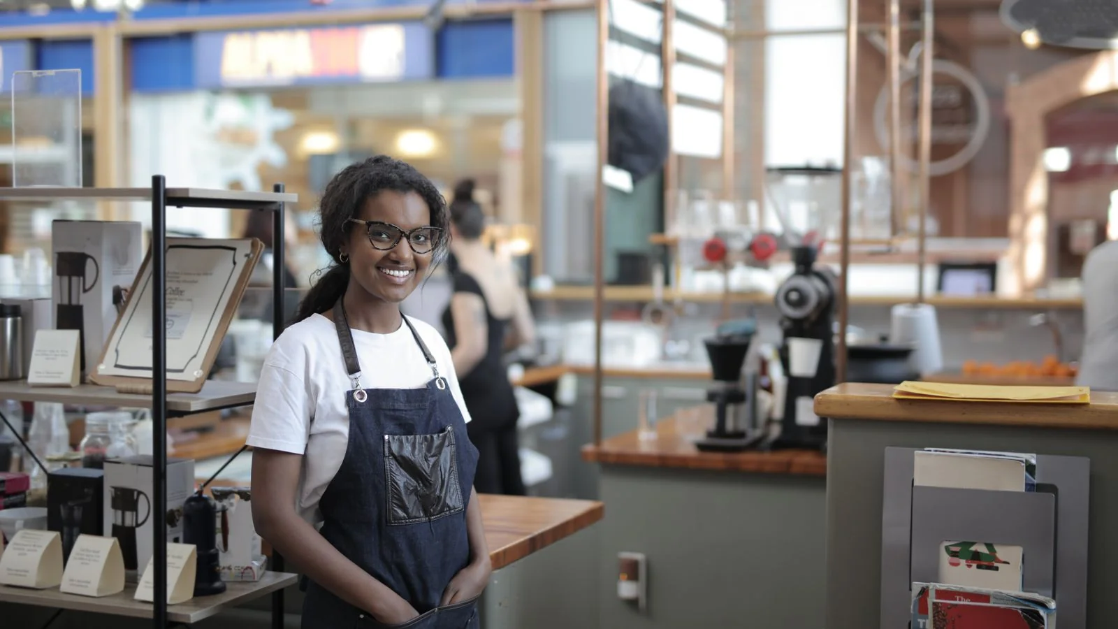 A smiling small-business owner in a work apron stands confidently in their shop.