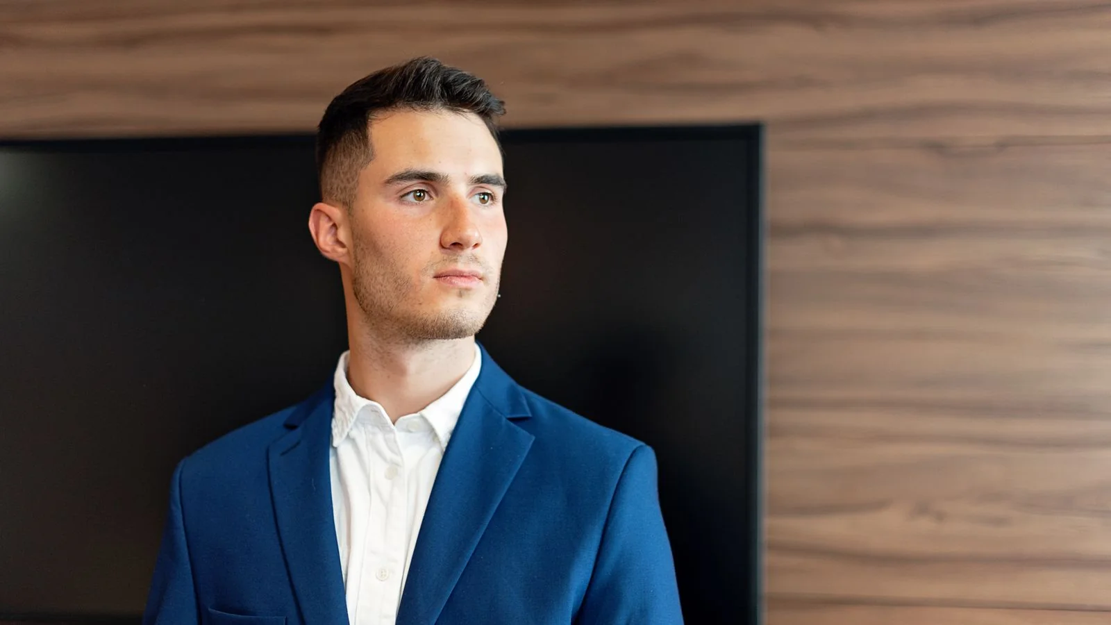 A confident young businessman in a blue suit stands in a modern office environment.