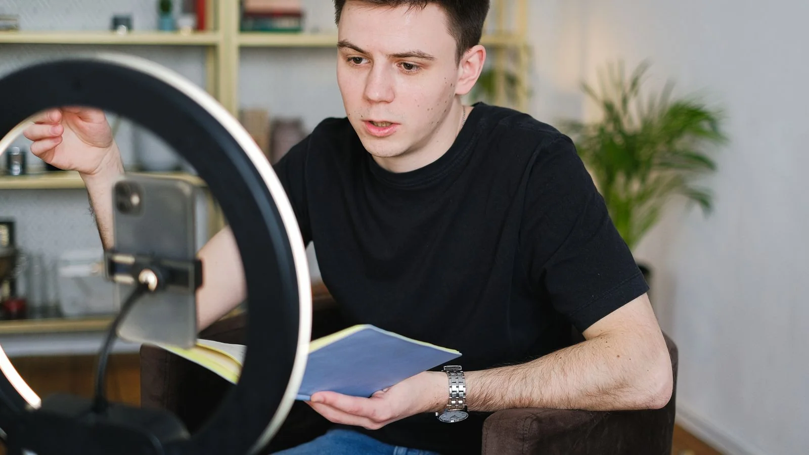 A man recording video content at home with a smartphone, ring light, and a notebook