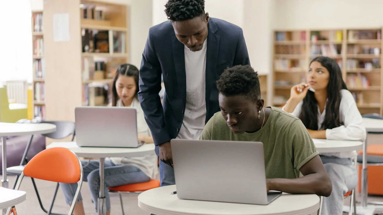 Students at a classroom desk listening to a teacher who is helping with their work on laptops