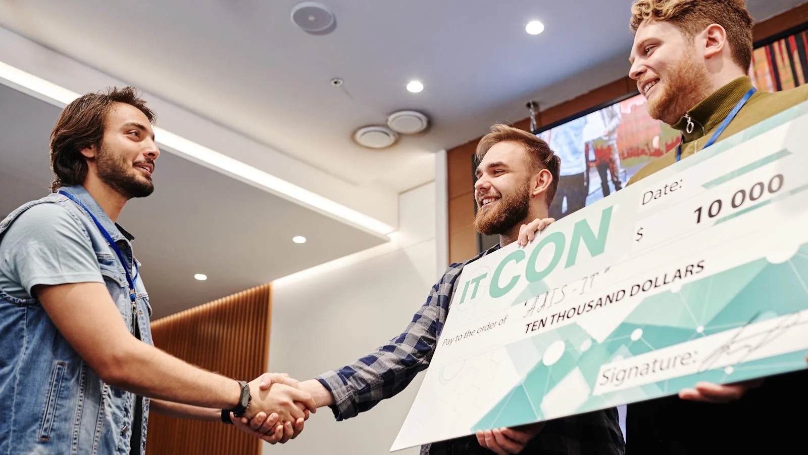 Two men celebrating with a large mock check, smiling and shaking hands at an indoor presentation ceremony