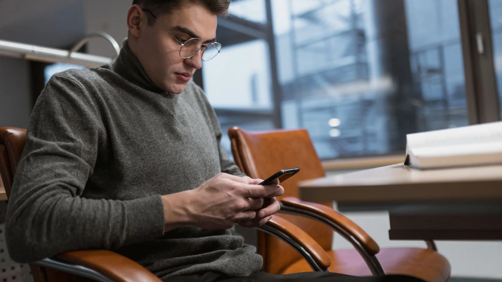 Young man in a grey sweater reading his smartphone at an office desk