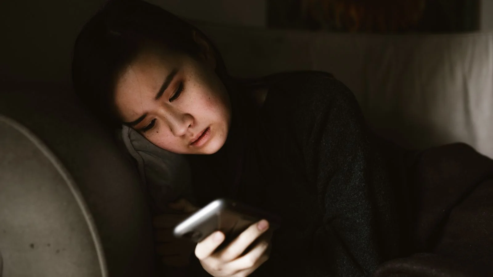 A person lying on a couch at night looking at a smartphone screen.