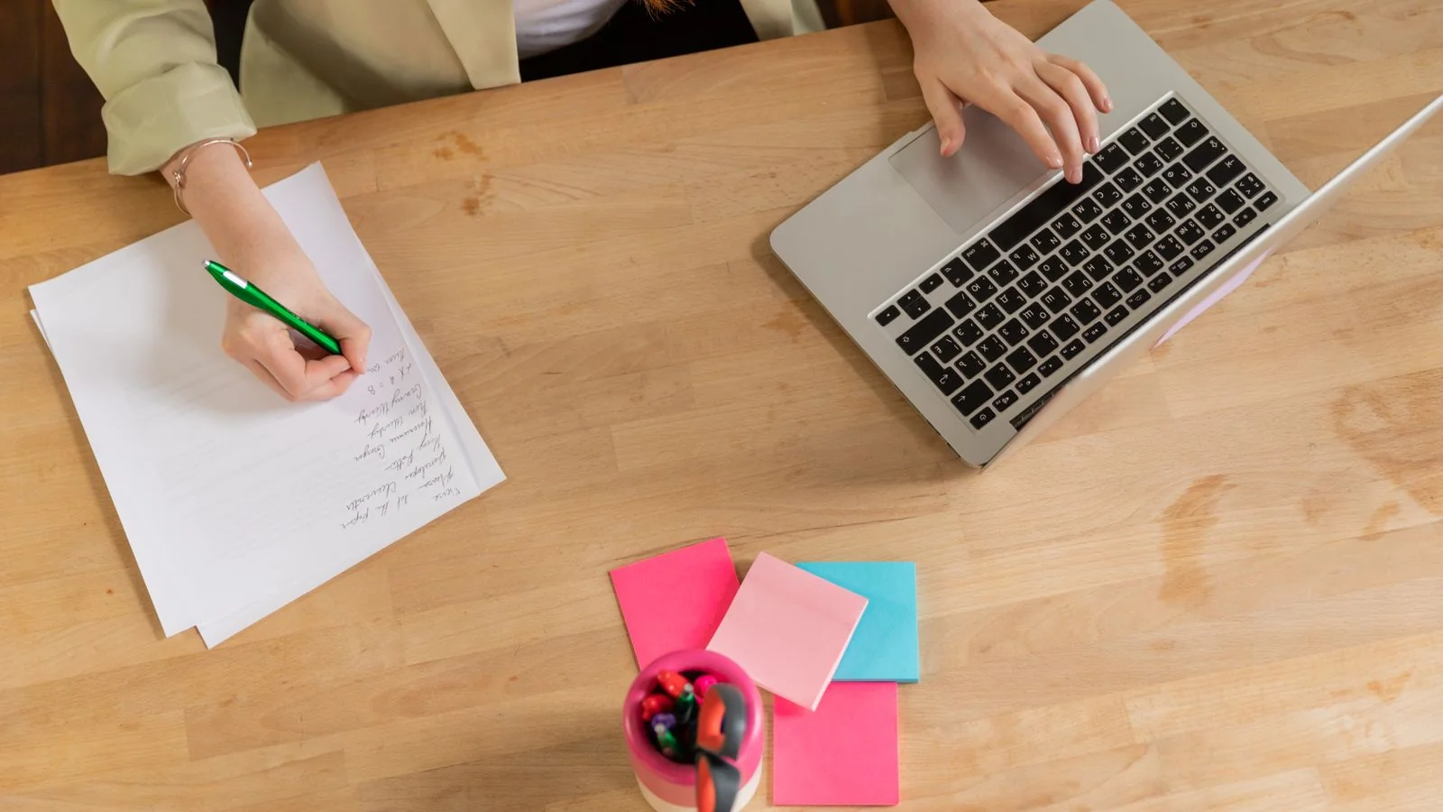 Overhead view of a person writing notes and typing on a laptop at a wooden desk