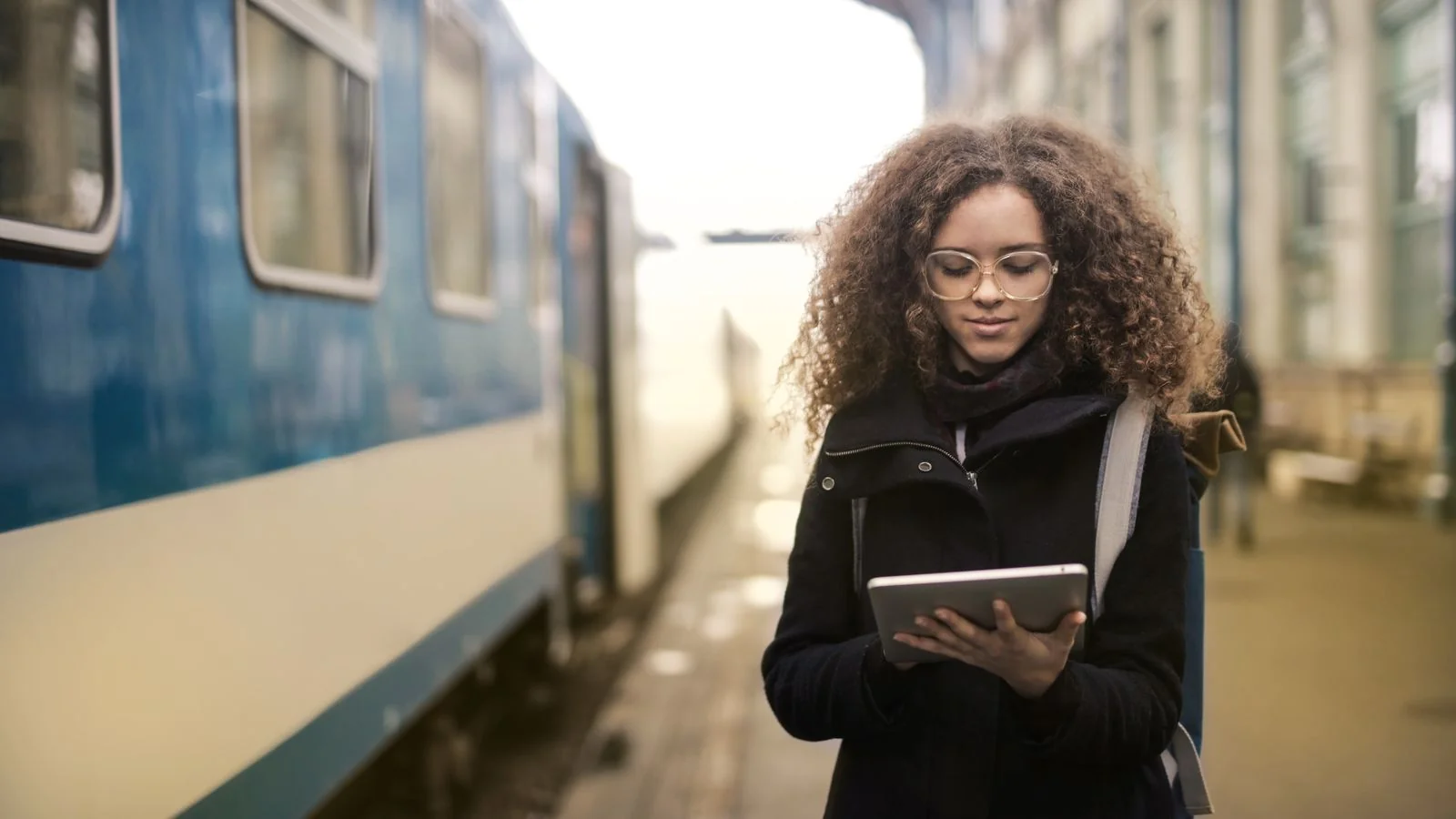 A young woman in a coat reading on a tablet at a train station platform